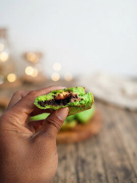 A close-up, high-resolution studio photograph of a dark-skinned hand holding a halved, vibrant green pandan-flavored mini pancake (known as Kue Cubit in Indonesia). 