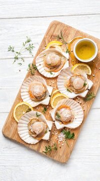 Fresh raw scallops in shells with lemon slices, herbs and sea salt on a wooden cutting board