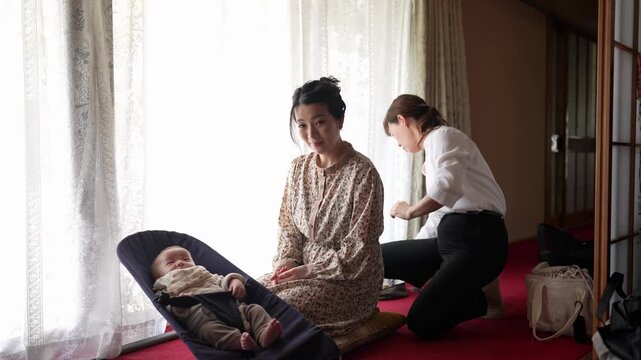 A seated woman waits calmly as another adult reaches for tools and continues preparing her hairstyle An infant remains visible beside her
