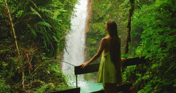 Woman watching tall waterfall cascading into bright turquoise pool below in Costa Rica