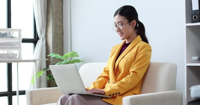 Young asian business woman focusing on her work, smiling at a laptop while sitting at a desk with documents and office supplies in a contemporary workspace