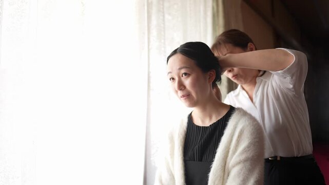 A woman looks forward in bright window light as another adult adjusts her hair from behind The scene captures quiet personal preparation