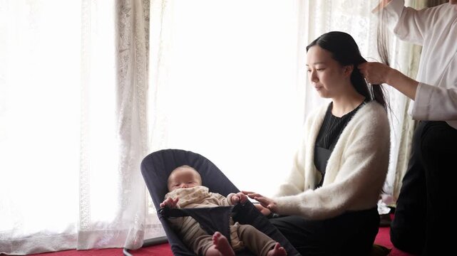 An adult smooths and aligns the hair at the crown of a seated woman head The infant beside her remains relaxed in a seat