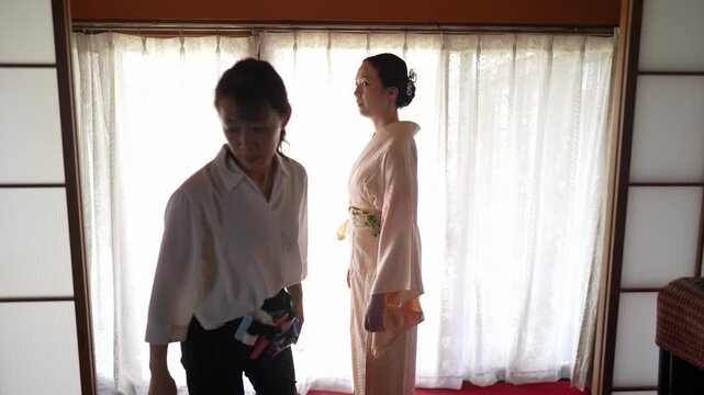A woman looks toward her clothing while a dresser steps aside after adjusting the pale kimono layers Bright curtains frame the scene