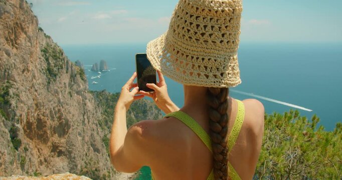Woman in straw hat photographing the sea from a cliff on Capri Italy with Faraglioni rocks in view