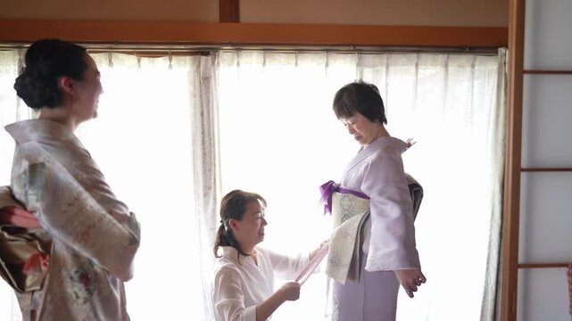An adult adds and tightens a purple decorative cord over a patterned obi Another woman in a floral kimono watches from nearby