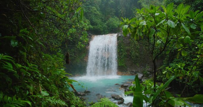Cascading waterfall rushes down rock into bright turquoise pool surrounded by lush jungle during wet rainy season