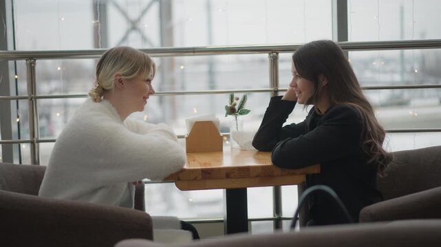 Two young women sit face to face at round wooden cafe table sharing warm conversation, gentle smiles, attentive gestures, soft backlight, urban window view, calm modern interior, with women