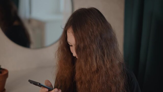 Young woman seated indoors uses smartphone with gentle scrolling and tapping gestures, head slightly tilted, relaxed shoulders, warm diffuse light, calm private moment and natural home setting