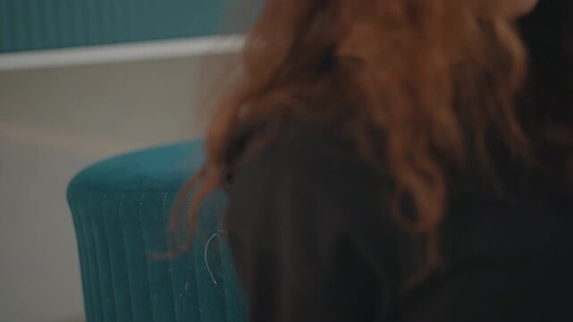 Close-up portrait of young woman with long curly hair seated beside teal upholstered chair, holding paper while reading aloud and reacting with focused gaze, open mouth, and calm indoor presence