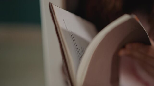 Close view of young woman reading with downward gaze while gently flipping thin paperback pages; fingertip supports inner page edge, visible curl, soft motion blur, and warm indoor light create calm