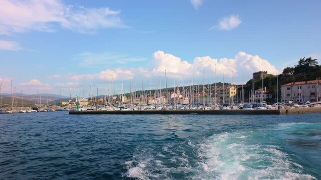 Scenic coastal adriatic marina of Muggia in Italy with sailboats, calm blue water, a foamy boat wake, waterfront buildings, green hills, and a bright summer sky