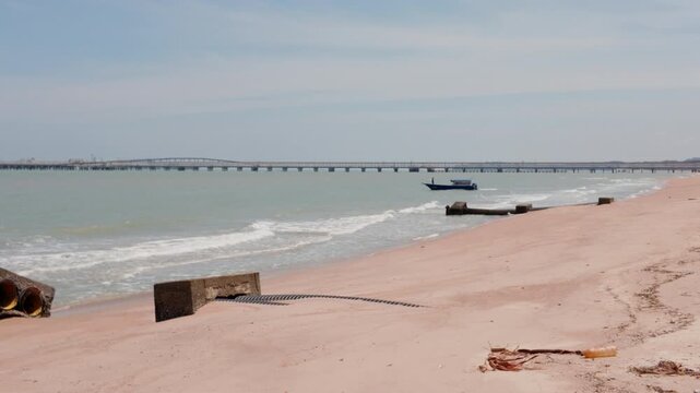 4K slow-motion static shot of Pantai Puteri beach with an outfall pipe, distant figure, and Melaka Energy Park refinery over the Strait of Malacca, Malaysia.