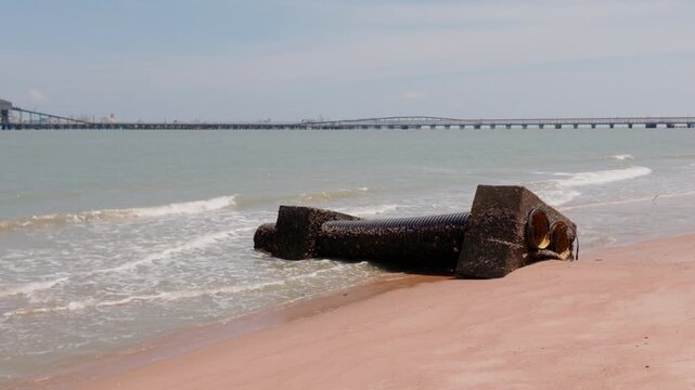 4K slow-motion static close-up of a weathered concrete outfall pipe on Pantai Puteri beach with gentle waves and the distant Melaka Energy Park refinery, Malaysia.