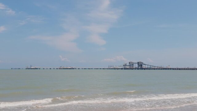 Zoomed static shot of the Melaka Energy Park refinery complex and pier over the Strait of Malacca, viewed from Pantai Puteri beach in Malaysia. 4K slow-motion static telephoto shot.