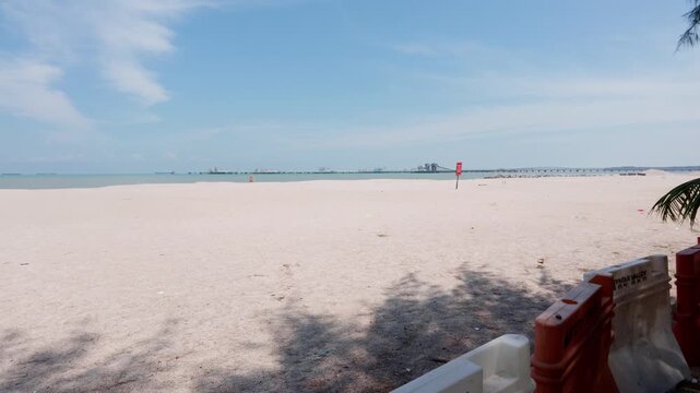 4K slow-motion static shot of Pantai Puteri's closed coastline behind safety barriers, looking toward the Melaka Energy Park refinery across the Strait of Malacca.