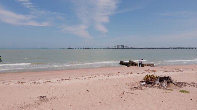 4K slow-motion static shot of Pantai Puteri's closed coastline with safety barriers, overlooking the distant Melaka Energy Park refinery across the Strait of Malacca.