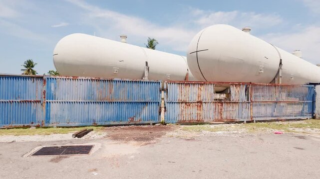 POV from a moving car driving past large white industrial pressure vessels and a rusted blue metal fence along Pantai Puteri in Melaka, Malaysia. 4K slow-motion POV driving shot.