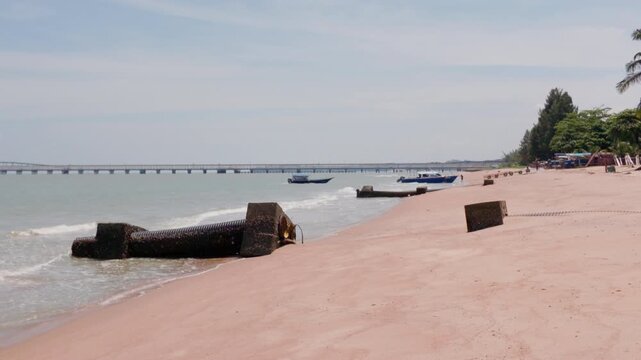 4K slow-motion static shot of Pantai Puteri beach with coastal outfall pipes, anchored boats in shallow water, and Melaka Energy Park refinery over the Strait of Malacca, Malaysia.