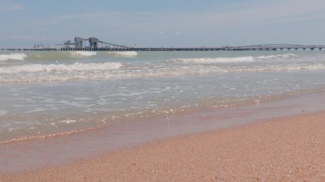 4K slow-motion close-up of gentle waves on Pantai Puteri beach, with the distant Melaka Energy Park refinery on the Strait of Malacca horizon, Malaysia.