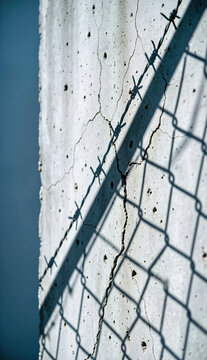 Vertical shot of a cracked concrete wall with barbed wire and chainlink fence shadows. Security barrier in bright daylight. Concept of restriction and confinement