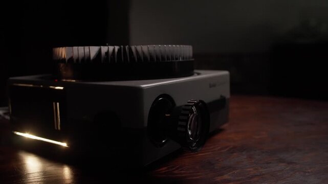 Vintage slide projector with illuminated lens rests on a dark wooden tabletop indoors, medium angled static as image displays in distance