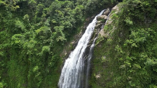 Dynamic aerial tilt-down tracking shot descending alongside Metaponto waterfall. Drone slides down parallel to the rushing water, showcasing the vertical cliff wall and surrounding jungle.