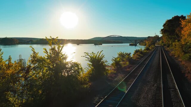 Railroad tracks follow the river toward an arch bridge in autumn near Ozark, Arkansas, aerial approach above glistening water