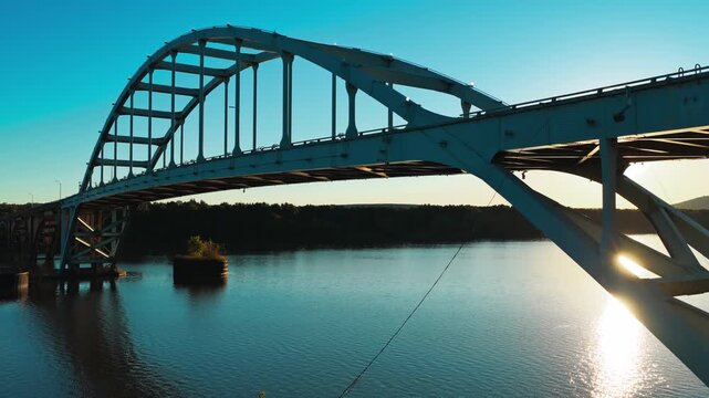 White steel arch bridge rises above a sunlit river in Ozark, Arkansas, aerial tracking angled pullback with glistening sunset glow, low upward angle
