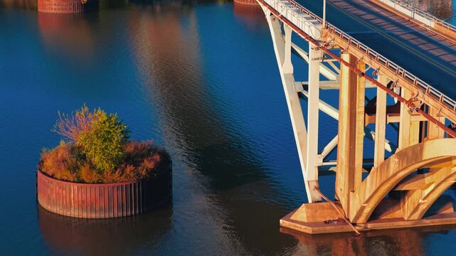 Bridge support and tree covered circular pier rise from Ozark, Arkansas river, aerial abstract pullback, rusted glow across curved arch