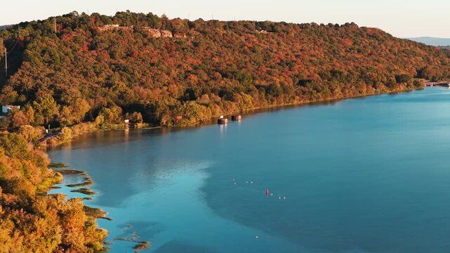 Blue river curves past dense autumn woodland near Ozark, Arkansas, aerial tracking right reveals dam at sunset