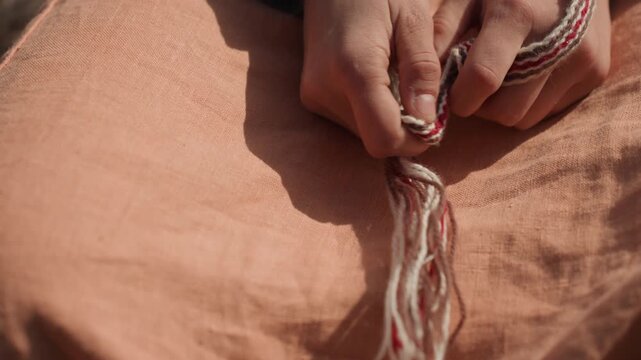 A close-up shot of hands meticulously tying a knot on sandy outdoor terrain, showcasing concentrated precision and care under natural light. This DIY and crafting scene is outdoor.