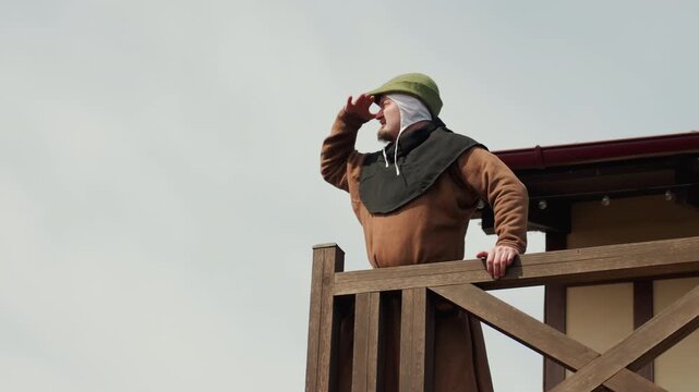 Mature bearded man wearing metal helmet and padded brown coat stands on elevated wooden lookout platform, gripping railing while shielding eyes and scanning distant horizon with alert focus