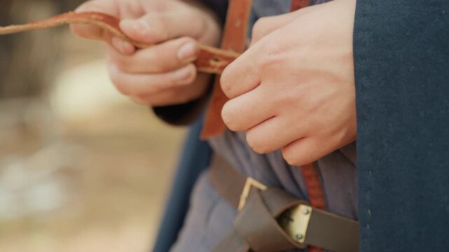 Close-up shows hands actively fastening worn leather strap and button over layered quilted garment beneath dark cloak, with fingers adjusting tension, securing closure, and revealing aged stitching