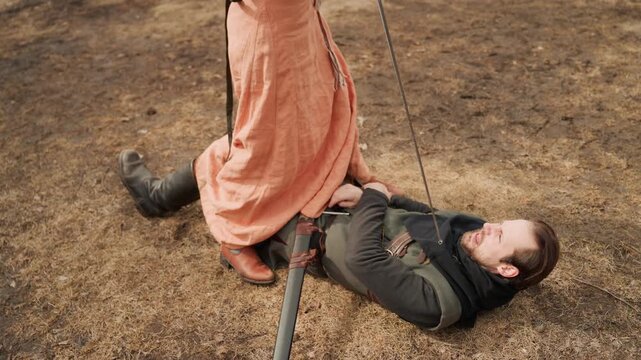 Young man lies face up on dry brown grass while another person stands over him, pressing boot into chest and holding strap; strong downward pressure, tense limbs, and rural setting create forceful