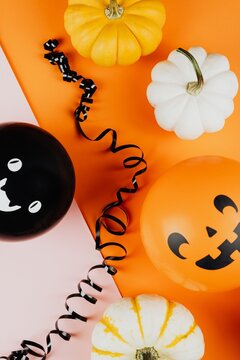 Halloween flat lay with yellow and white mini pumpkins, black curling ribbon, and ghost/jack-o'-lantern balloons on split pink and orange background &mdash; top view with copy space