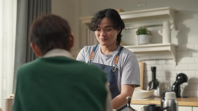 Man in denim apron smiling while brewing pour-over coffee, teaching friend at home kitchen counter, coffee beans and drip equipment on table, weekend hobby and hospitality concept, warm morning light.