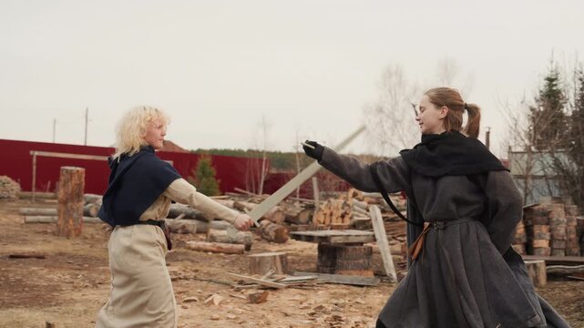 Two women in period costume move together across rustic outdoor yard after staged sword fight, dark-cloaked woman guiding lighter-dressed companion with gentle touch while capes, skirts, and swords