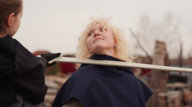 Curly blond youth leans back with tilted head and upward gaze while companion holds wooden staff across shoulders, creating visible tension, controlled movement, and engaged playful confrontation