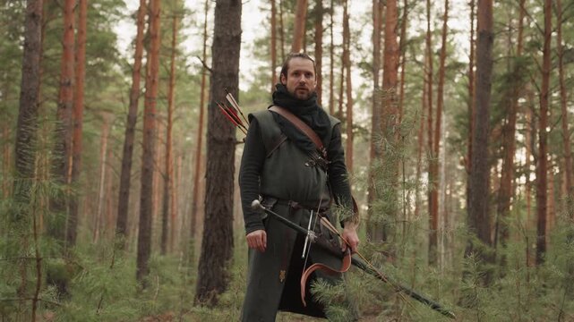 Bearded man in earth-tone outdoor clothing walks through pine forest, holding longbow at hip and carrying quiver of arrows on back; forward step, steady torso, upward focused gaze, with archer, man