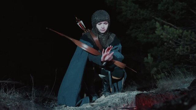 Young archer kneels in grassy forest clearing at night, drawing bow with arrow nocked and aimed ahead, focused expression, steady hands, and taut stance creating tense fantasy action under low light