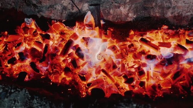 Close-up top-down view of rectangular fire pit filled with red-hot charcoal and glowing embers as vigorous flames rise and flicker across cracked coals, casting intense warm light, with ember, flame