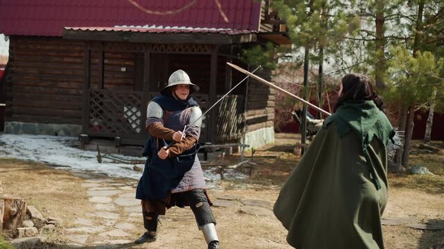 Costumed combatants face off in outdoor historical reenactment, armored fighter lunges with polearm while cloaked opponent recoils near rustic cabin, fence, snow and dry grass, with medieval, armor