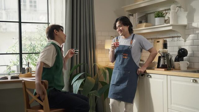Two men chatting and laughing while drinking coffee in cozy home kitchen, one wearing denim apron, morning routine and casual conversation, warm domestic lifestyle concept with natural light.