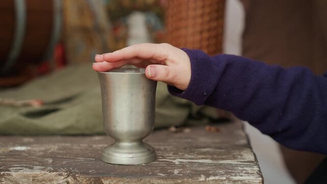 Close view of adult hand releasing coin into aged metal goblet on chipped wooden table, with brief rim contact, visible deposit gesture, rustic texture, soft indoor light, and shallow depth focus