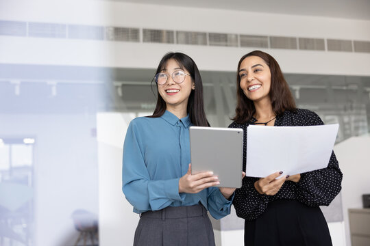 Two laughing businesswomen standing in office holding tablet and papers, look into distance, engaged in teamwork, documents discussion, working on joint task or online project. Career and achievements