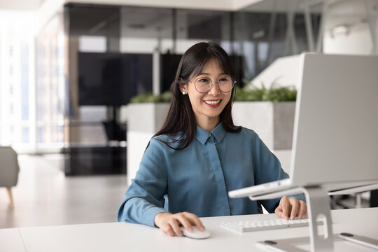 Smiling Asian woman, staff member working at ergonomically optimized desktop in office, reviewing report, preparing online presentation, feel satisfied with company position and comfortable workplace