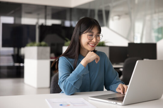 Smiling young Chinese professional woman, office employee using laptop sit at desk at contemporary workplace, reading received business proposal, lead correspondence to client, engages in online tasks