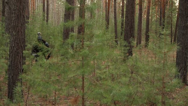 Person in dark clothing moves carefully through dense pine forest, showing forward stride, slight body turn, and cautious navigation between straight trunks in muted green woodland light