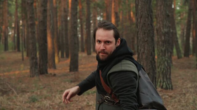 Bearded man in layered medieval clothing performs archery in autumn pine woodland, drawing wooden bow, releasing arrow with clear torso rotation, then turning toward camera while holding focused